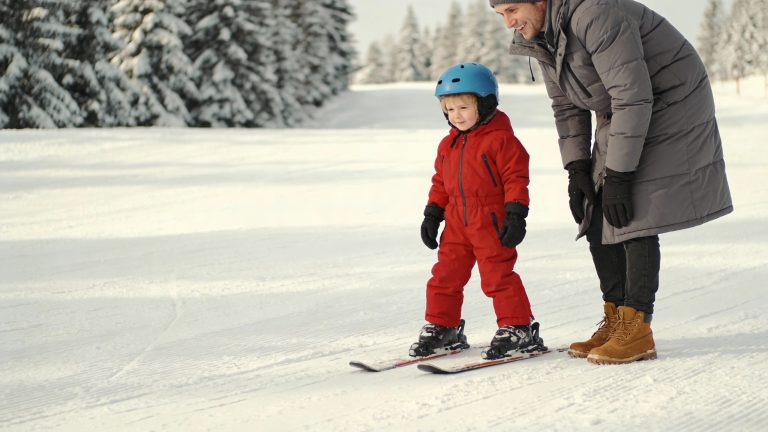 Parent and child on a gentle beginner ski slope, child in a red ski suit, both smiling, wide open bunny hill, soft winter sunlight.